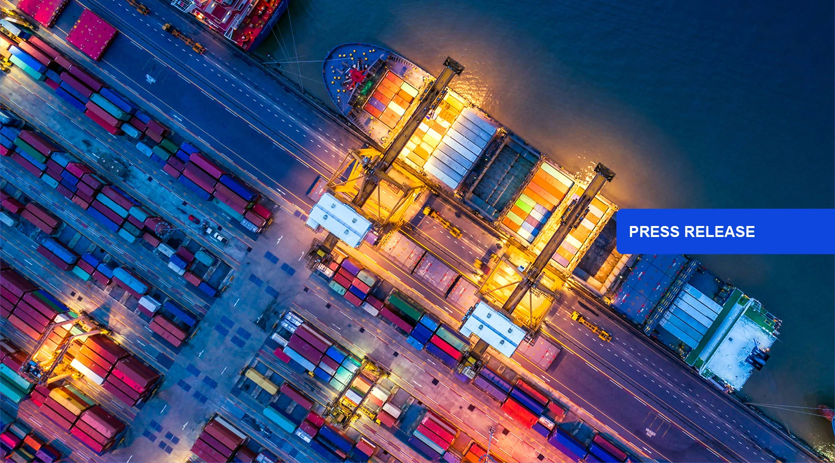Aerial view of a busy shipping port at night, with cargo ship and containers. Press Release.