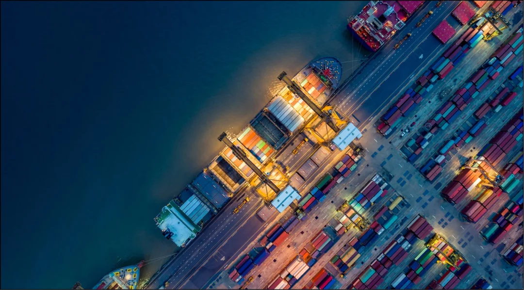 Aerial view of a busy, illuminated shipping port at night with cargo ships and containers.