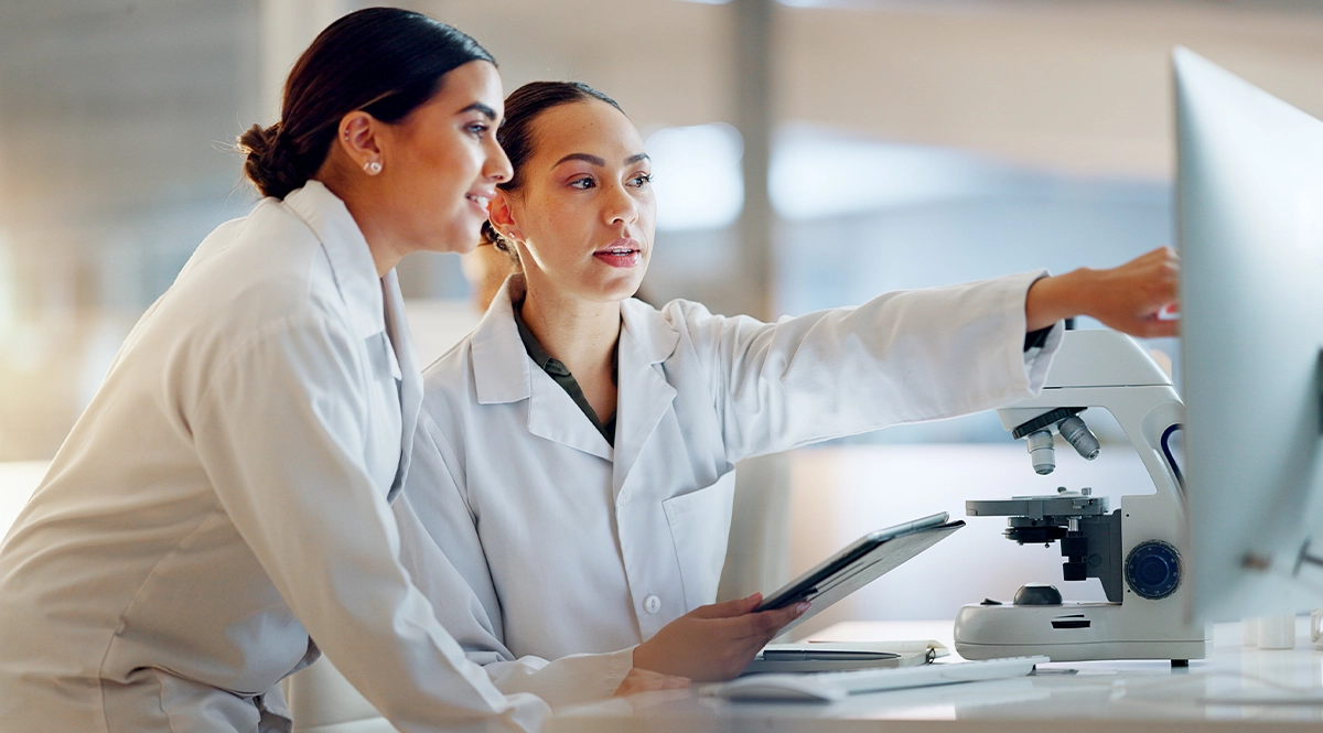 Scientists in lab coats review data on a computer and tablet next to a microscope.