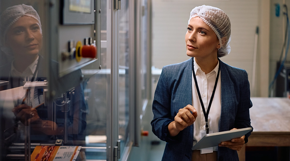 A food safety inspector wearing a hairnet and holding a tablet reviews equipment on a production floor.